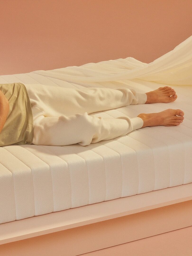 Person lying on a white mattress with salmon-colored lighting, wearing a green shirt and beige sweatpants.