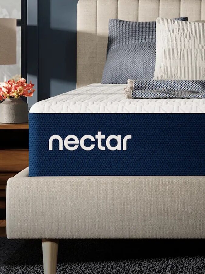 Close-up of a Nectar mattress on a beige bed frame with a textured headboard, surrounded by blue and gray bedding. A bedside table with a lamp and flowers is visible on the left.