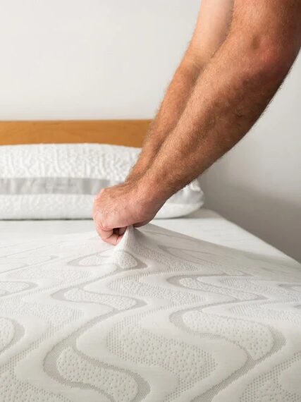 Person adjusting a mattress cover on a bed with two pillows against a wooden headboard.