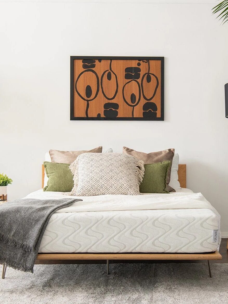 Minimalist bedroom with a wooden bed, white and green bedding, abstract wall art, and a floor lamp. Plants and a nightstand with a lamp are on the sides. "365 Night Sleep Trial" text is visible.