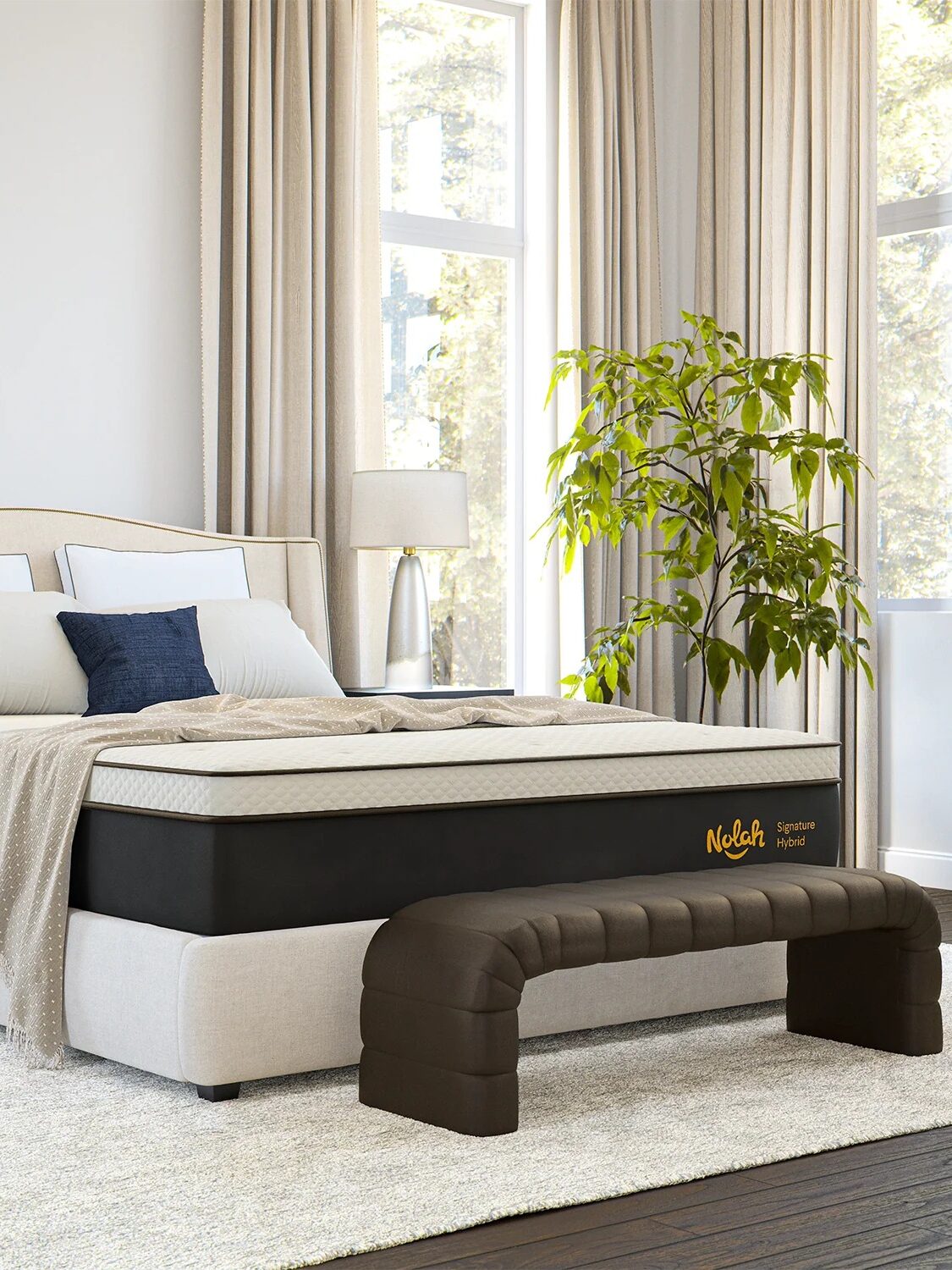 Bedroom with a neatly made bed, a black and white mattress, bedside tables with lamps, and a potted plant by the window. A padded bench is at the foot of the bed.