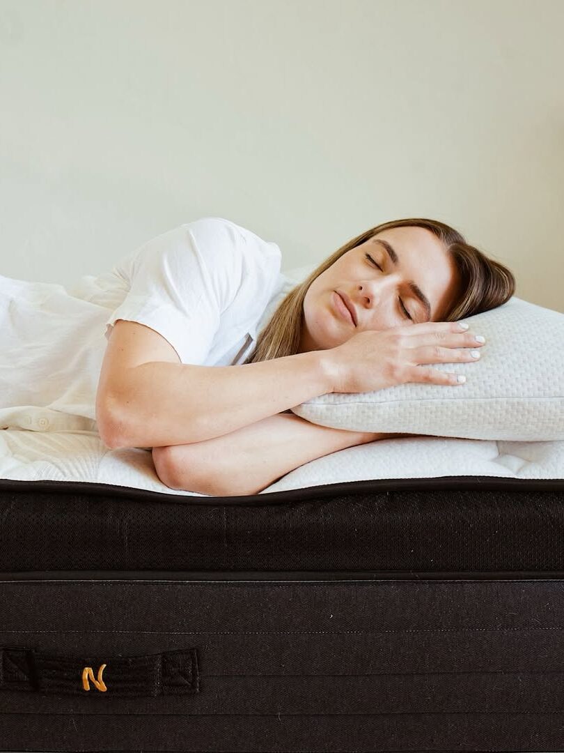 A person in a white outfit sleeps on a mattress, resting their head on a pillow, with eyes closed and facing sideways.