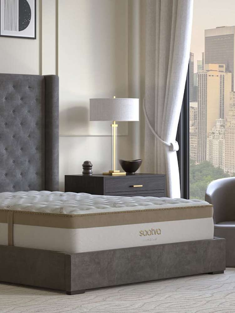 Bedroom featuring a large gray tufted bed with a plush mattress, flanked by two dark wooden nightstands with lamps, against a backdrop of cityscape through a window.