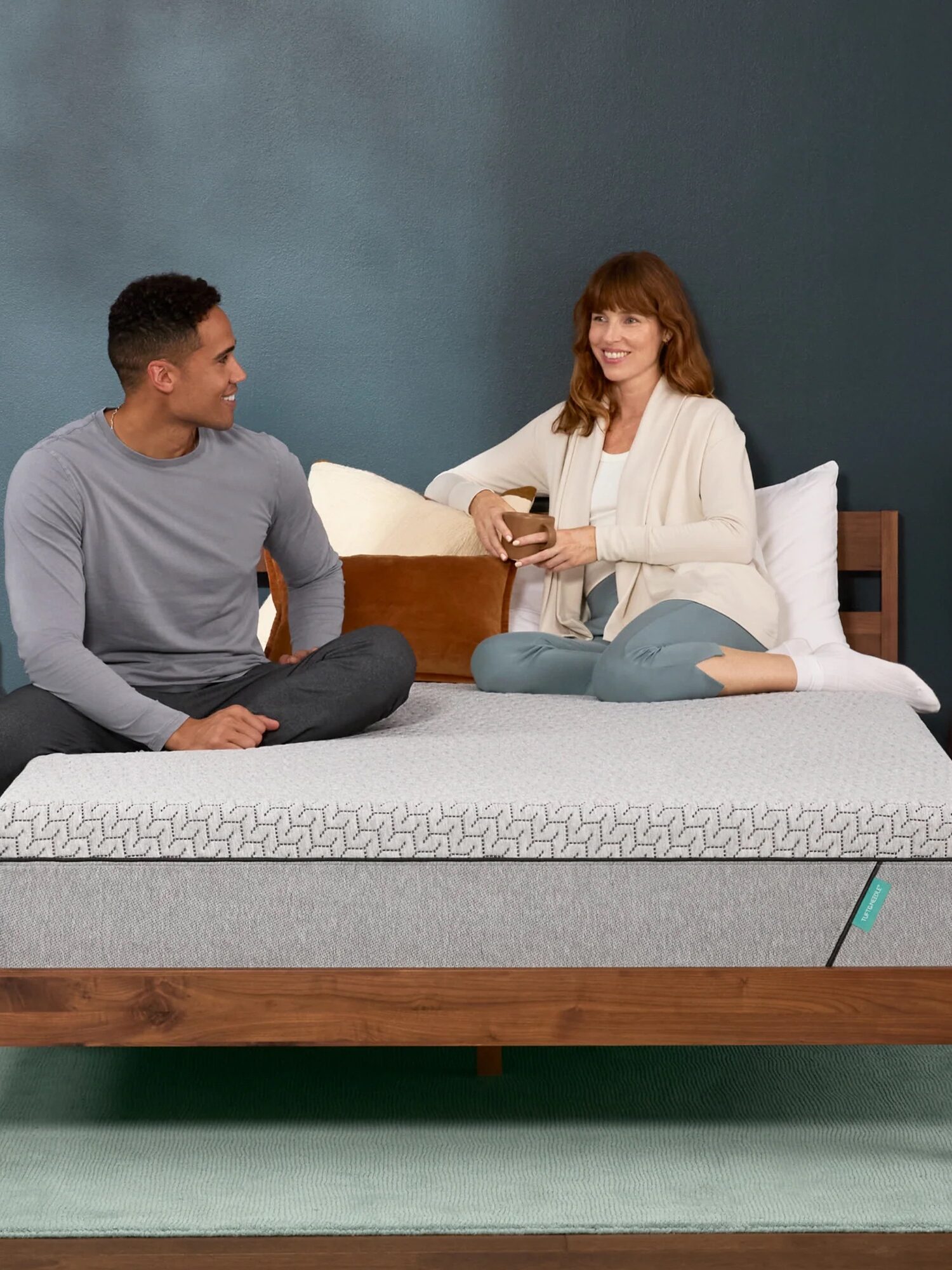 A man and a woman sit on a modern bed with a patterned mattress and wooden frame. They appear to be talking and smiling. A lamp and a potted cactus are on wooden nightstands.