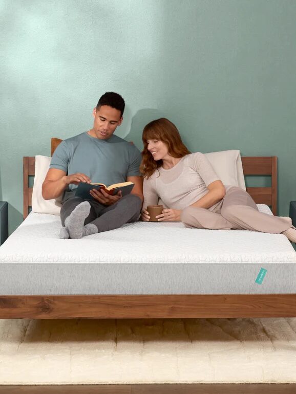 Couple sitting on a bed; the man reads a book, and the woman holds a mug, smiling. They are in a bedroom with green walls and modern decor.