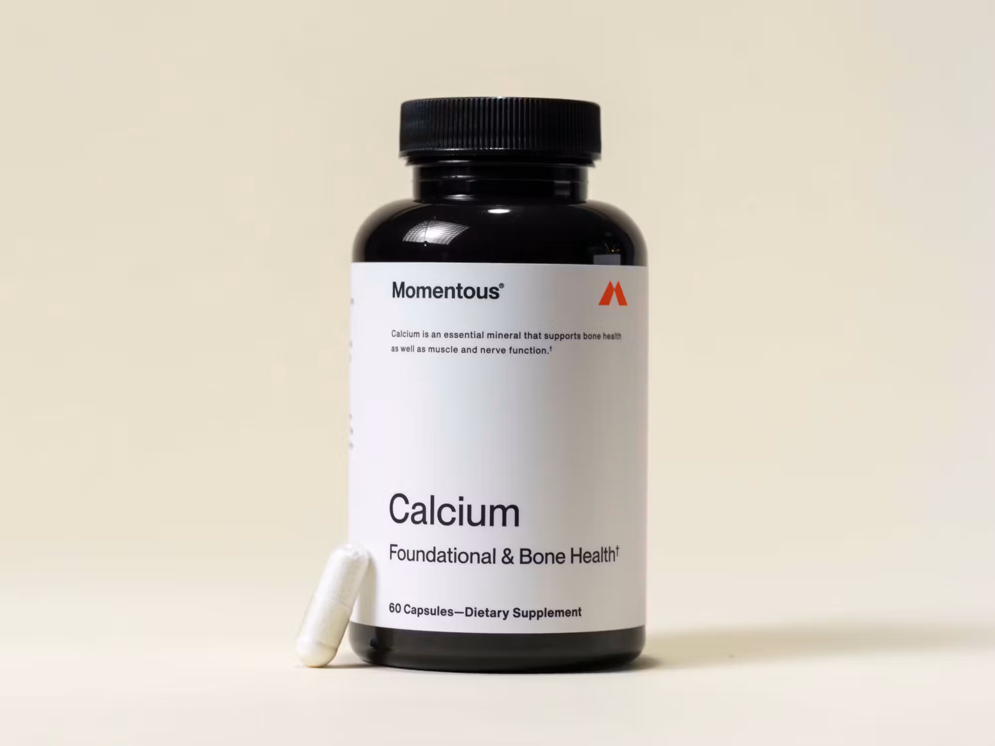 A black bottle labeled "Momentous Calcium Foundational & Bone Health" dietary supplement with one white capsule placed beside it on a beige surface.