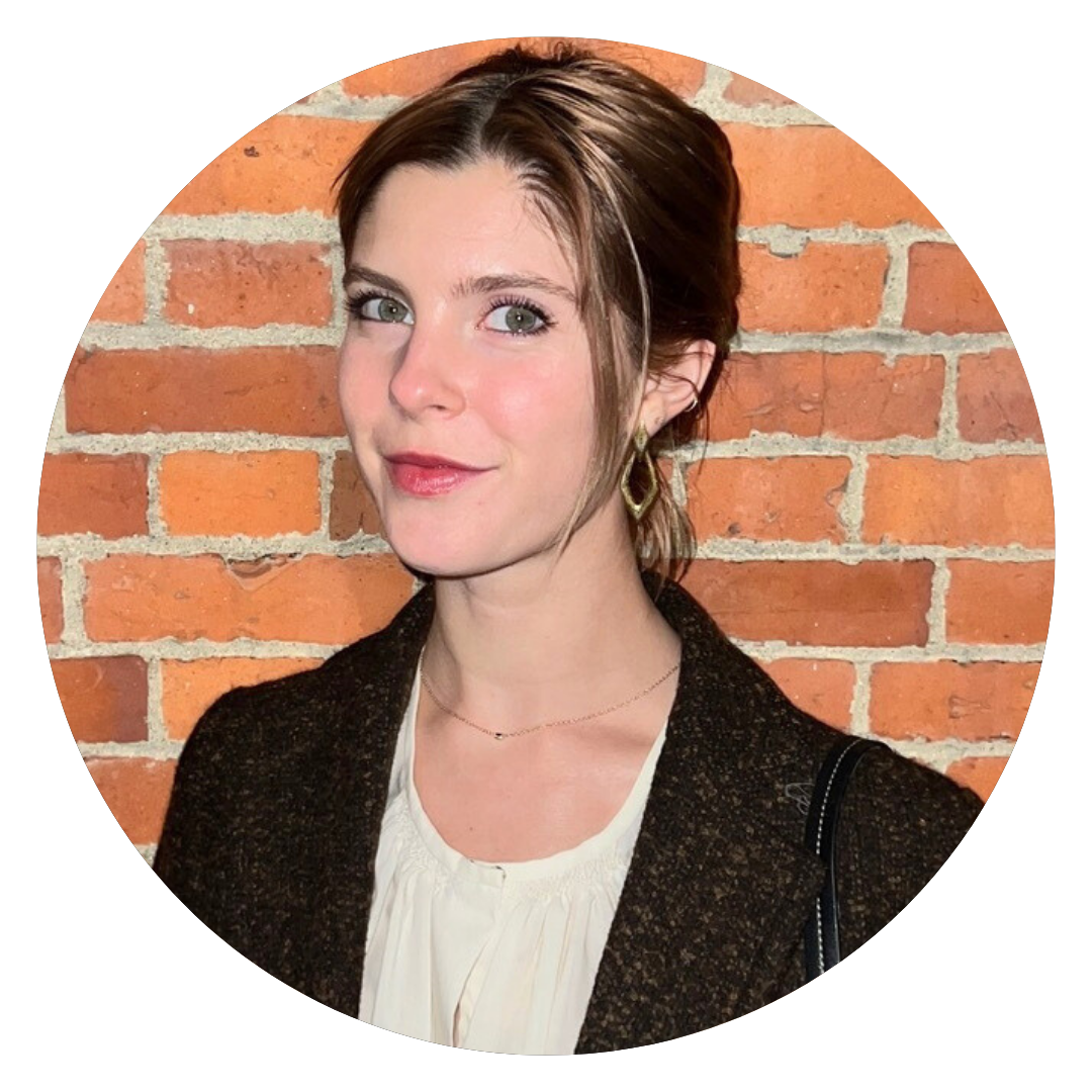 A woman with fair skin and brown hair is standing in front of a red brick wall, wearing a dark jacket over a light blouse and gold hoop earrings.