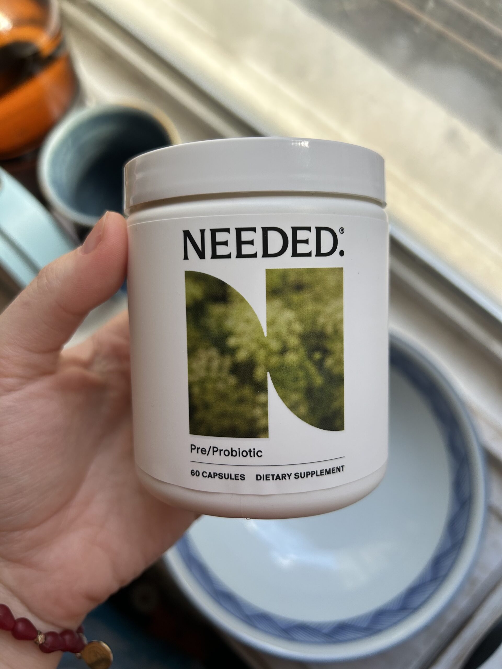 A hand holds a white container labeled "Needed Pre/Probiotic," containing 60 dietary supplement capsules, near a window with bowls in the background.