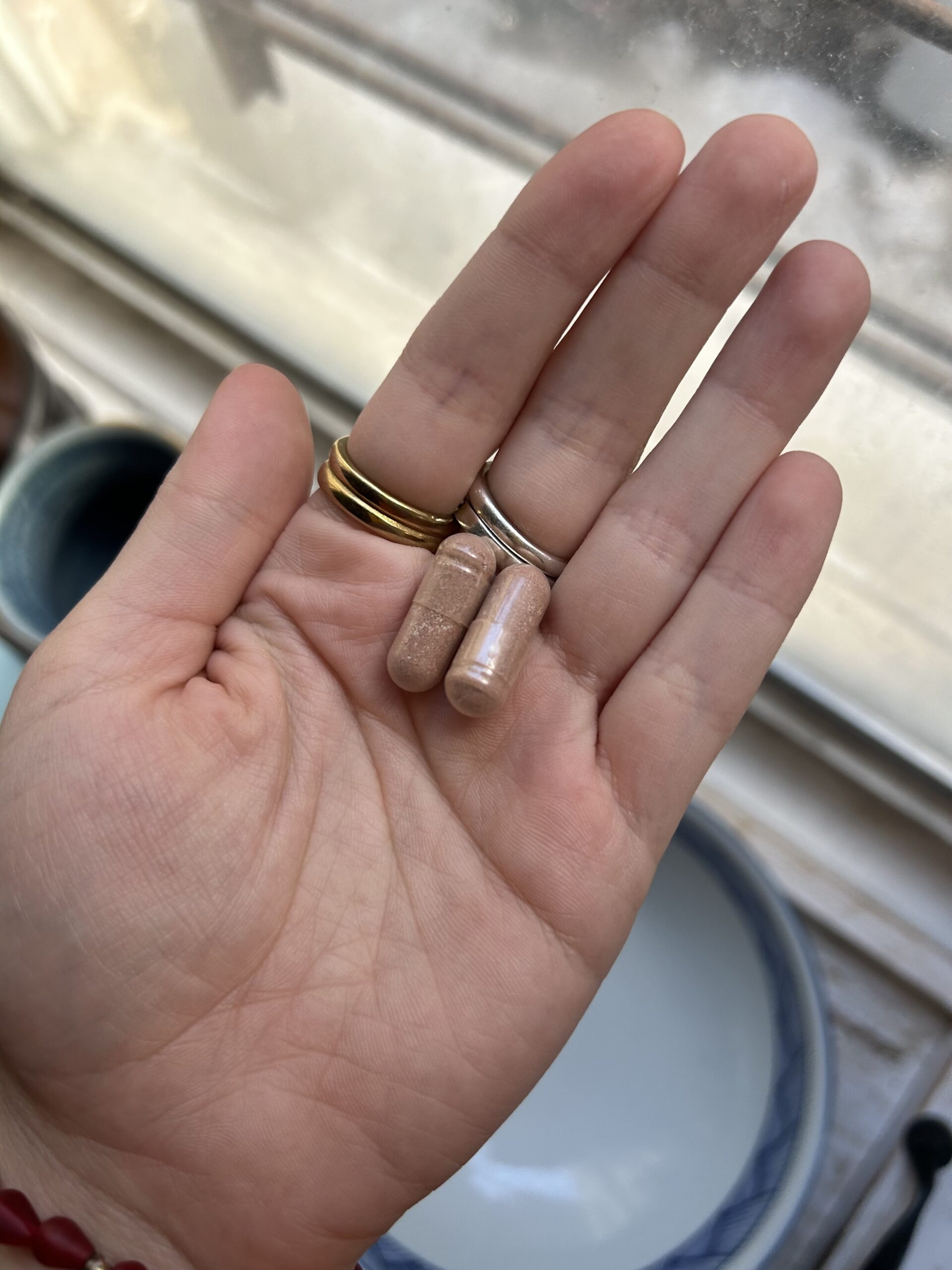 A hand with multiple rings holds two beige capsules above a white dish near a window.
