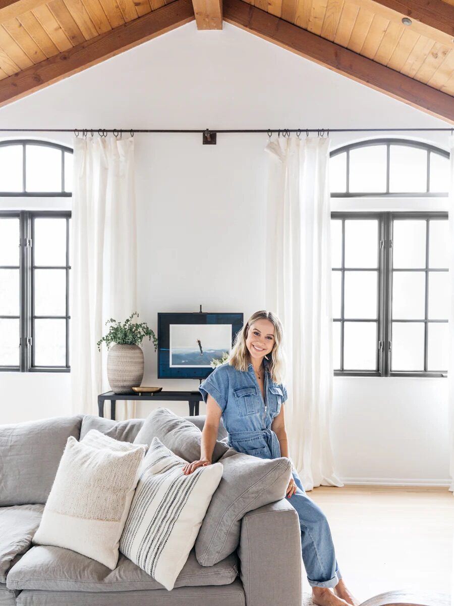 A woman in a blue jumpsuit sits on a gray couch in a bright living room with large windows, wood ceiling, and white curtains. A framed picture and a potted plant are on a table behind her.