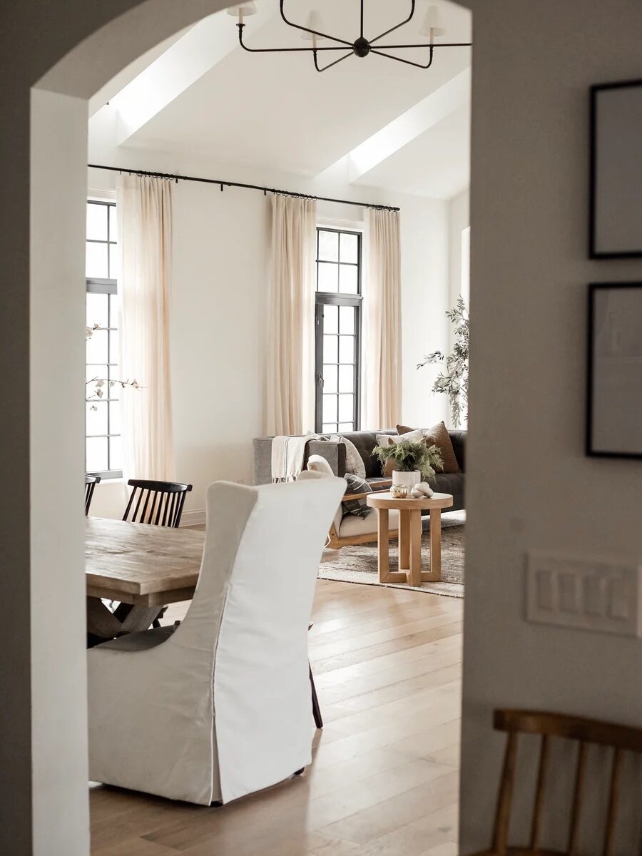 View of a modern living and dining room through an archway, featuring white walls, large windows with sheer curtains, wooden dining table, and a seating area with a sofa and coffee table.