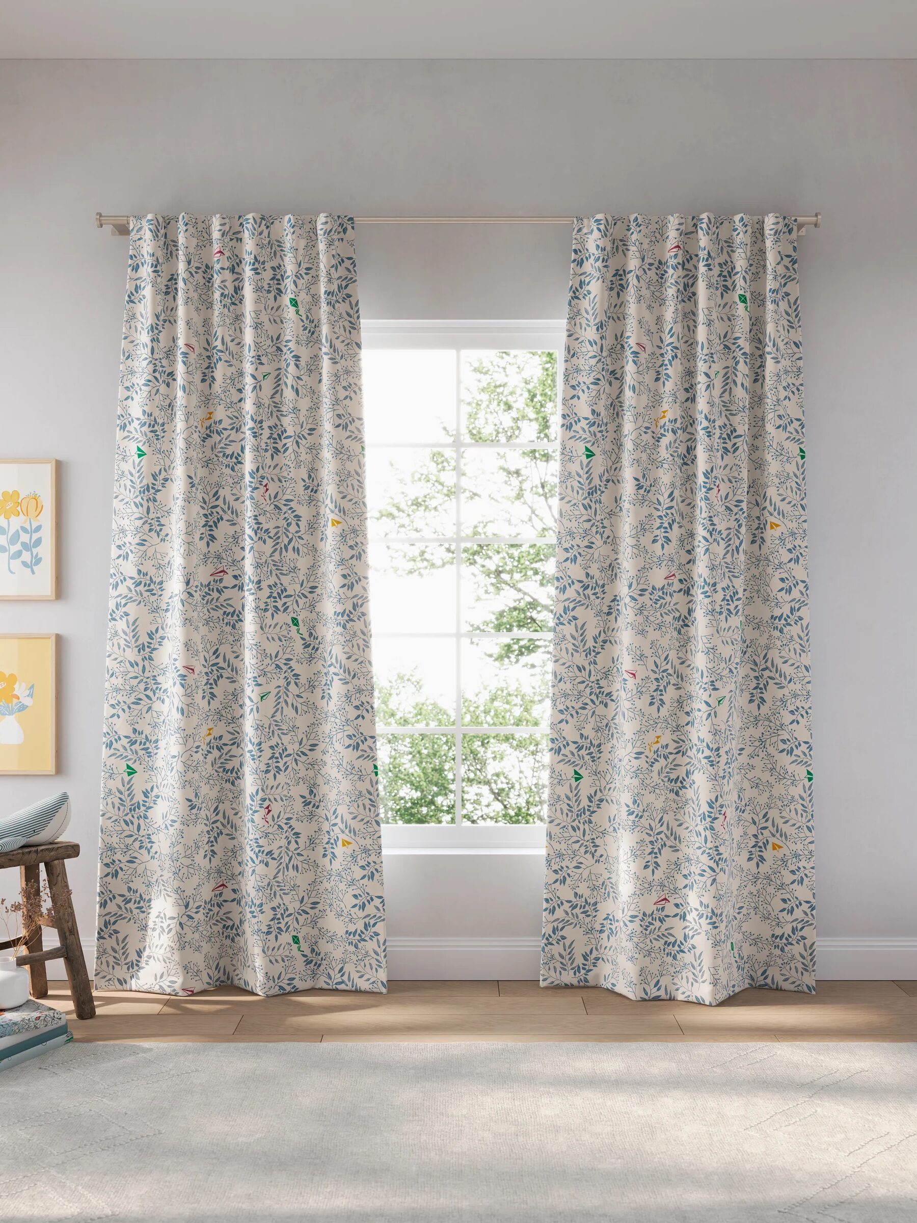 Floral curtains hang in front of a window, with a sunny view of trees outside. Framed flower art and a chair are on either side.