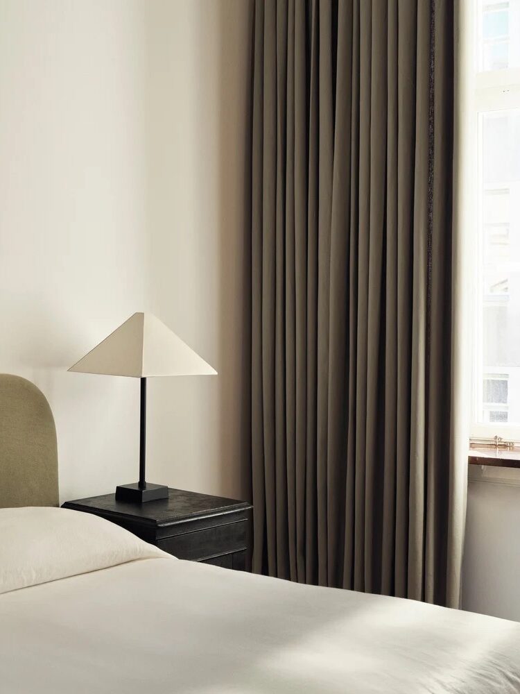 A minimalist bedroom with a bed, a dark nightstand, a geometric lamp, and floor-length curtains. Natural light enters through a window on the right.