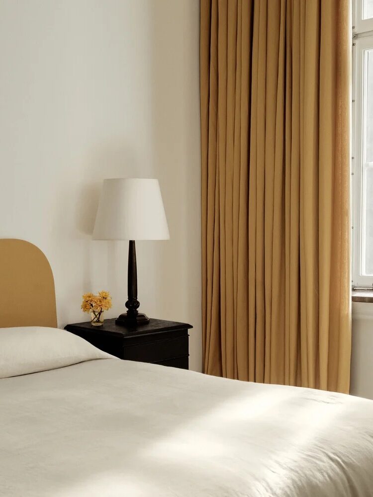 A bedroom with a beige bed, black nightstand, white lamp, and yellow curtains. Sunlight filters through the window, casting gentle shadows.