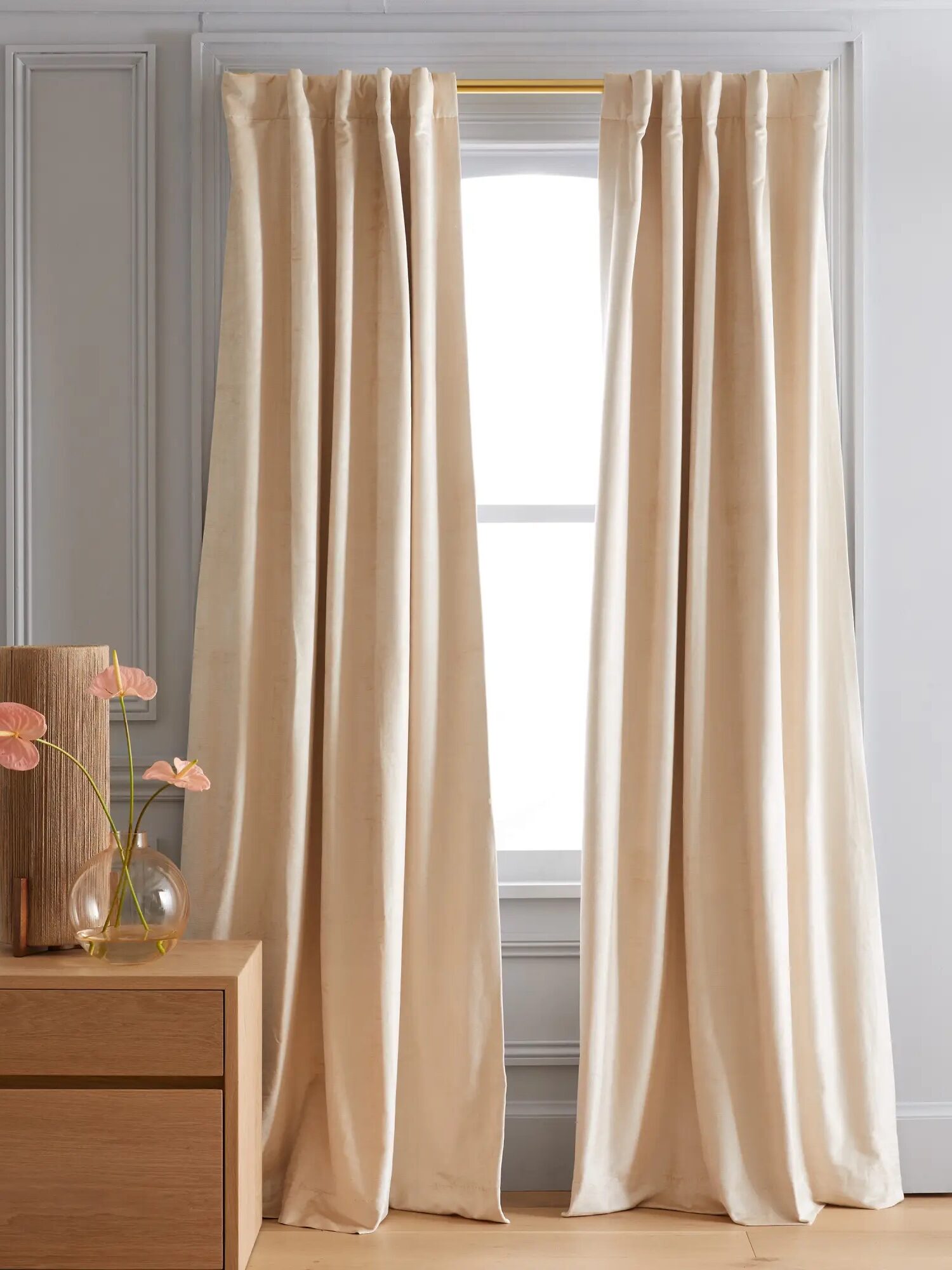 Cream-colored curtains hang from a rod in front of a window. A wooden dresser with two vases holding pink flowers is on the left side. The setting is in a room with light-colored walls.