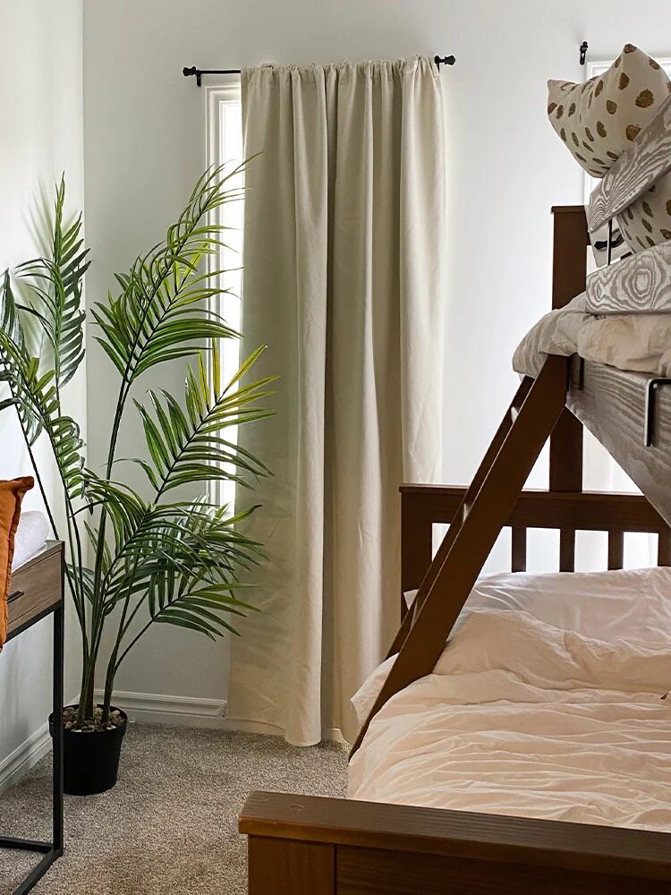 Bedroom with a wooden bunk bed, a potted palm, artwork with an animal, orange cushion, a bench, and a curtained window.