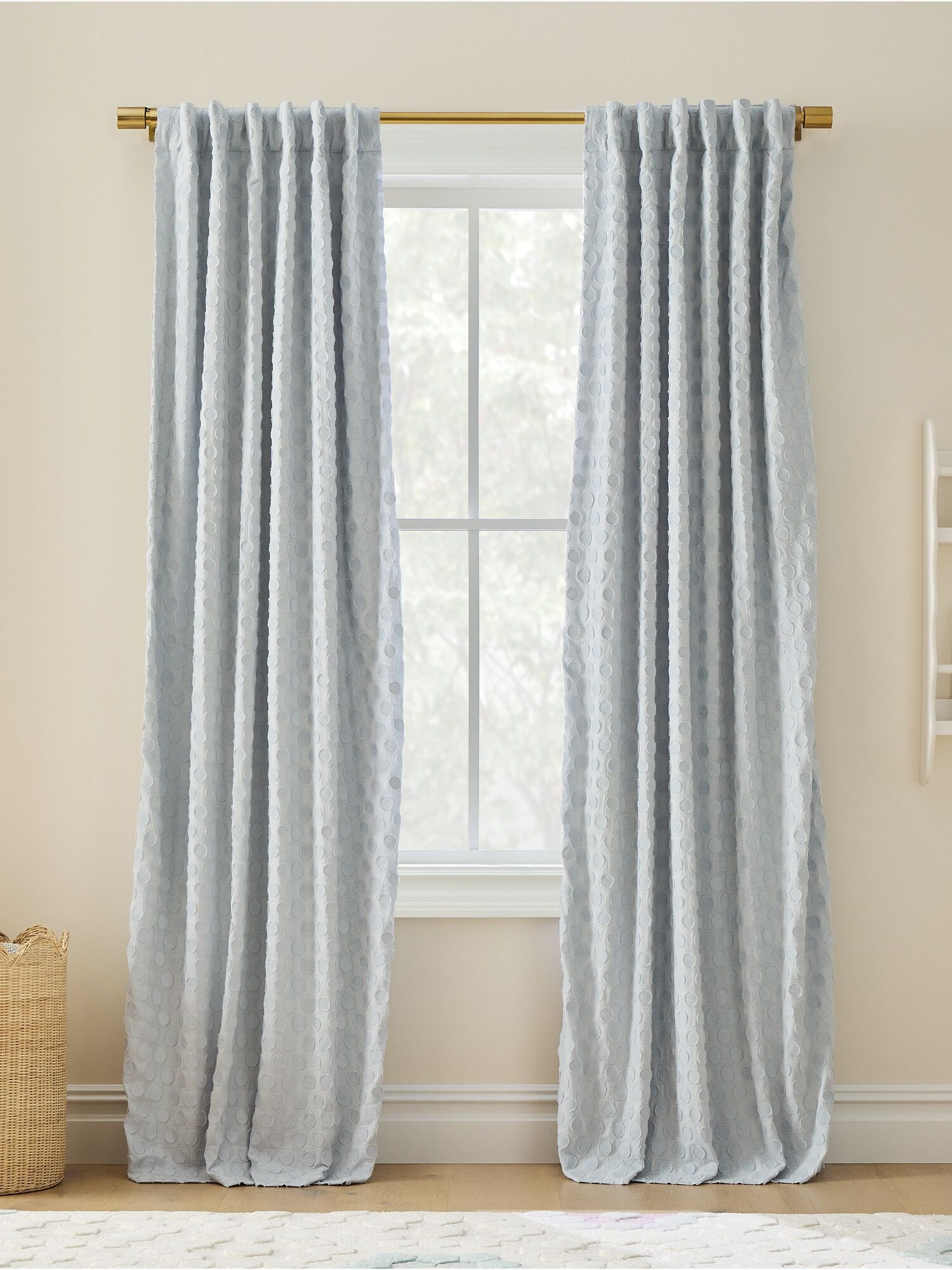 Light blue curtains hang on a gold rod over a window in a neutral room with a woven basket and wall shelves.