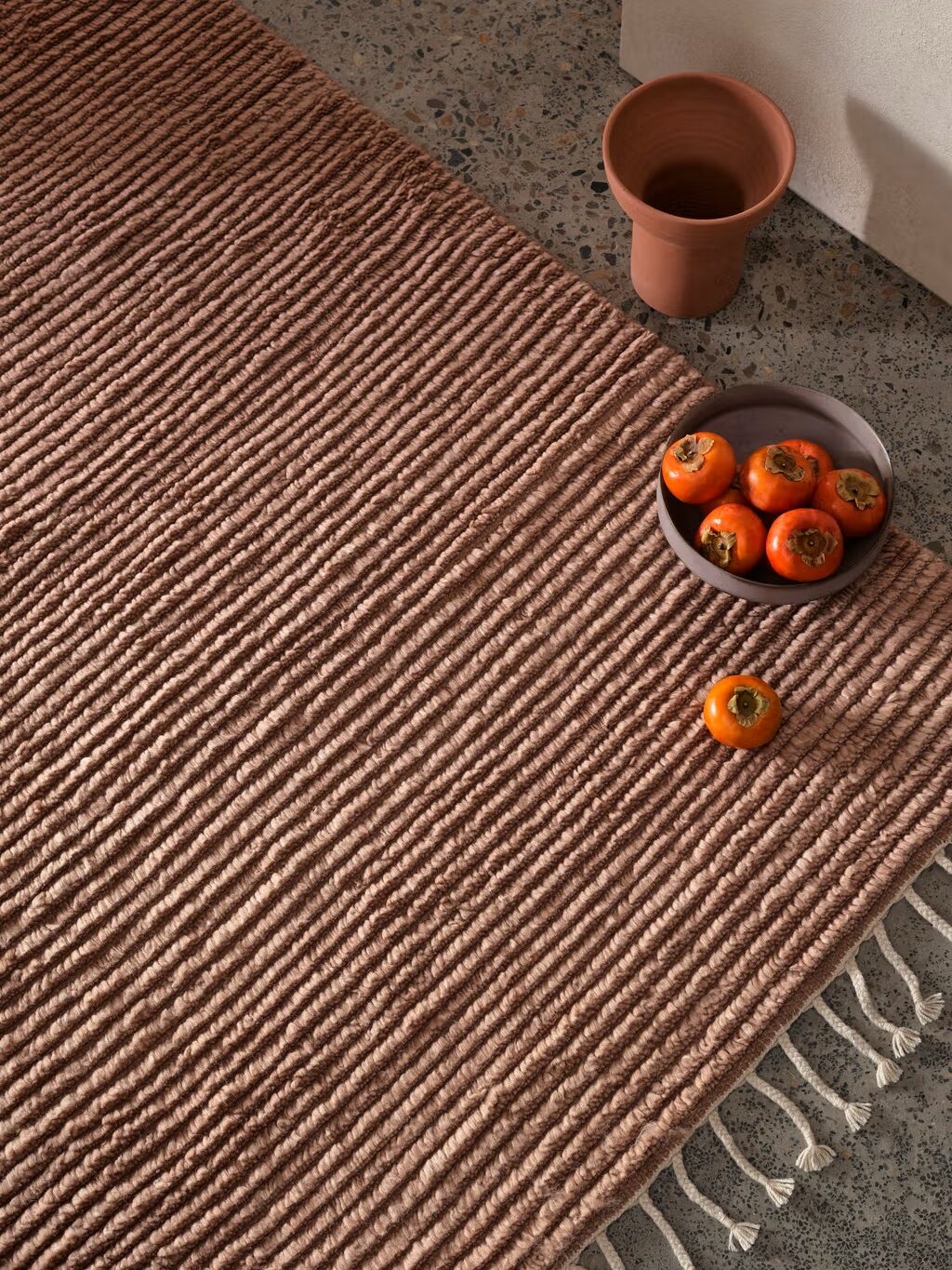 Brown textured rug on a stone floor with a fringed edge. A bowl containing five persimmons and a terracotta cup are placed on it.