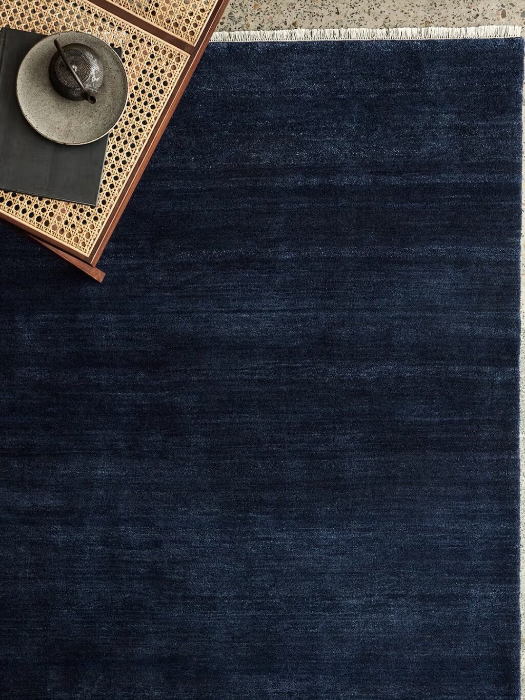 A dark blue rug on a floor with a corner of a rattan table and a gray plate with a small bowl on top.