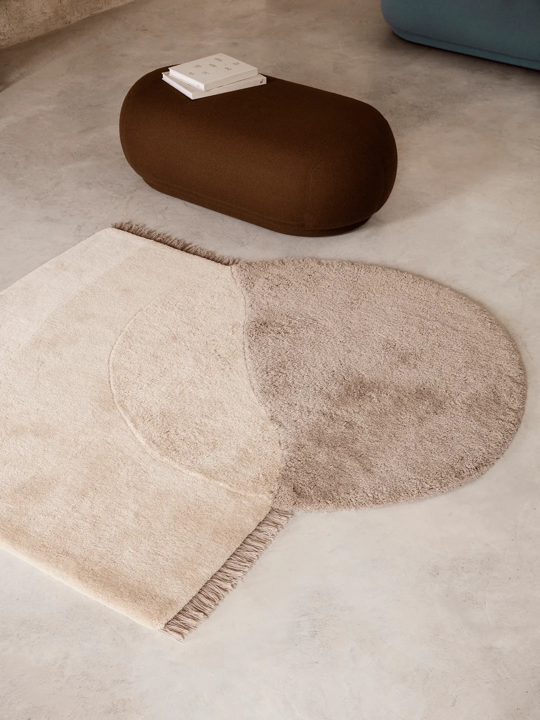 A geometric rug with overlapping beige and taupe sections on a concrete floor, near a brown oval ottoman with a book on top.