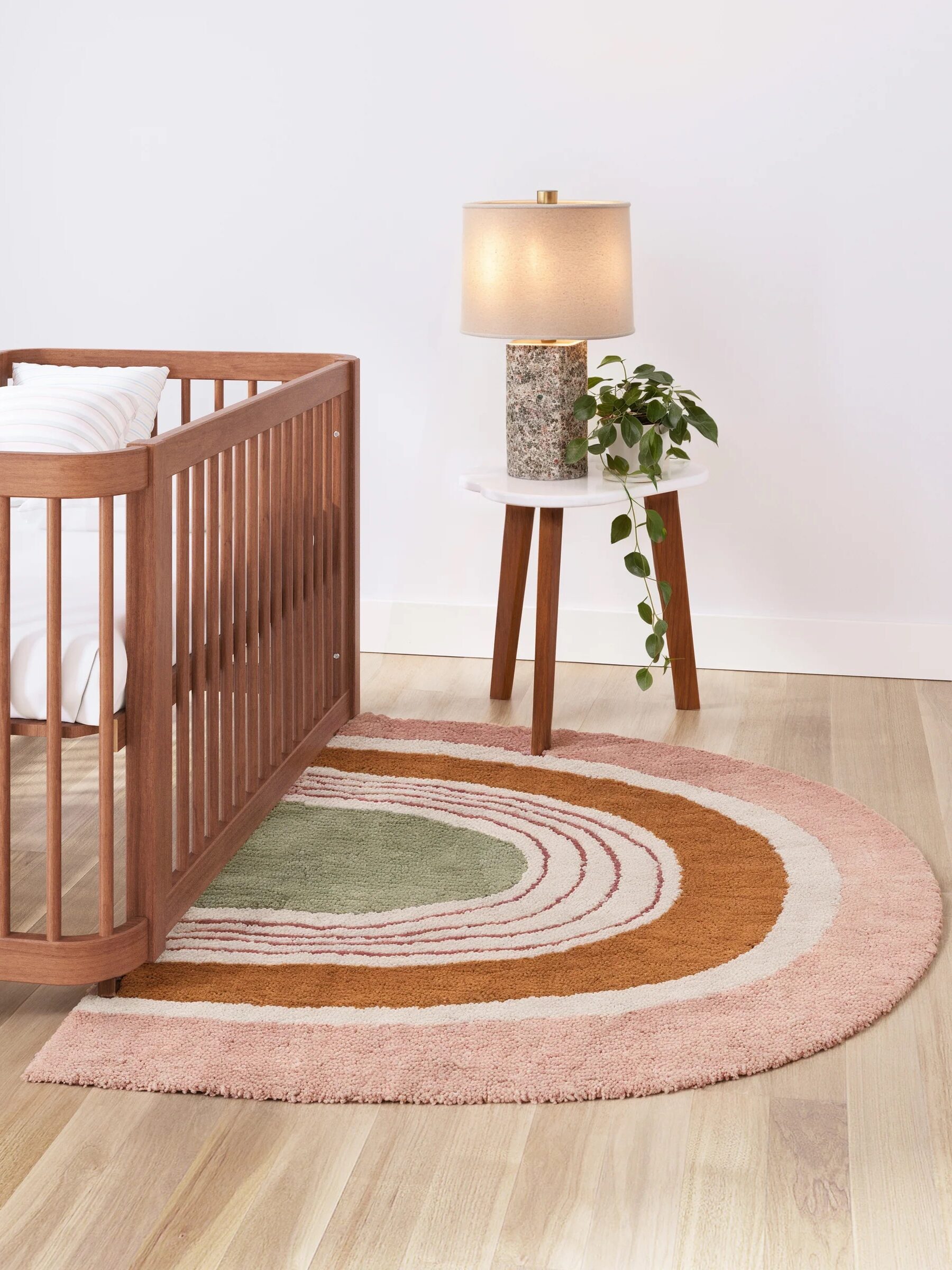 A nursery features a wooden crib, a round multicolored rug with curved patterns, and a small table with a lamp and potted plant on light wooden flooring.