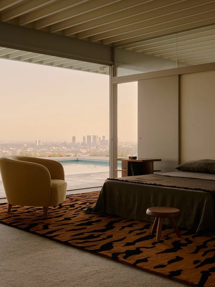 Modern bedroom with a large window view, minimalist decor, a yellow armchair, and a patterned rug. A cityscape and pool are visible outside.