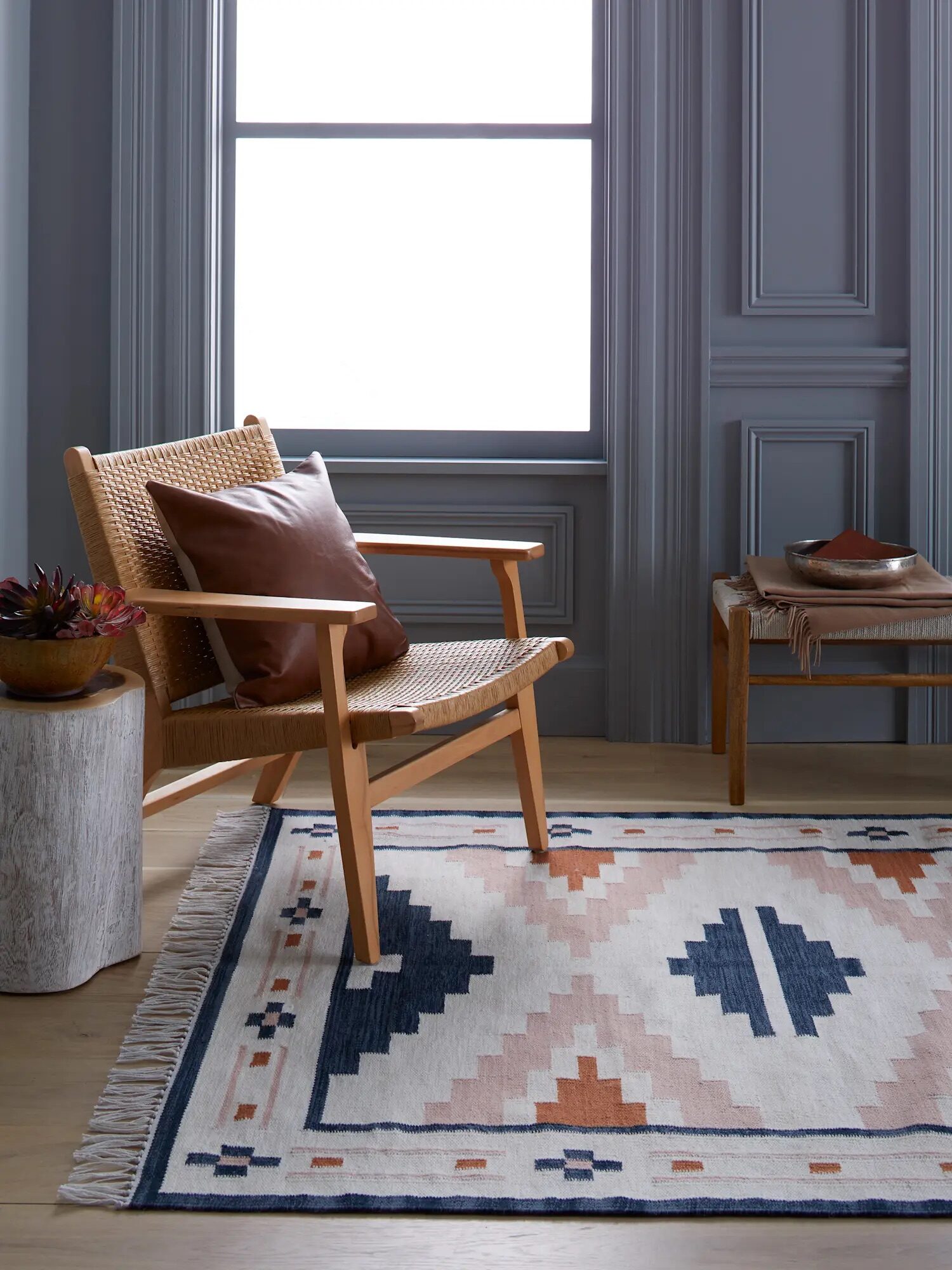 A cozy room features a wooden chair with a leather cushion, a small bench with decor items, and a patterned rug on a wooden floor, with two large windows in the background.