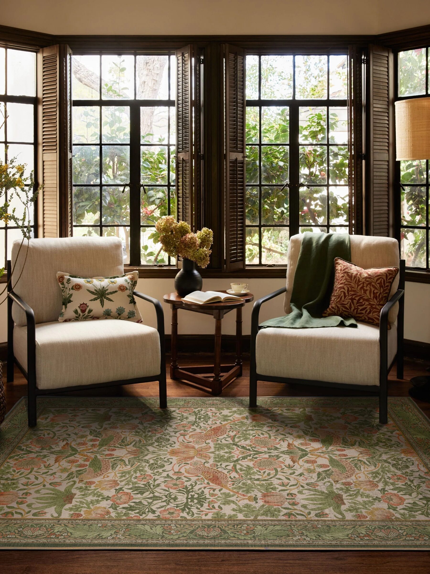 Two armchairs with patterned cushions face a small round table in front of large windows. A floral rug covers the floor, and a lamp stands in the corner.