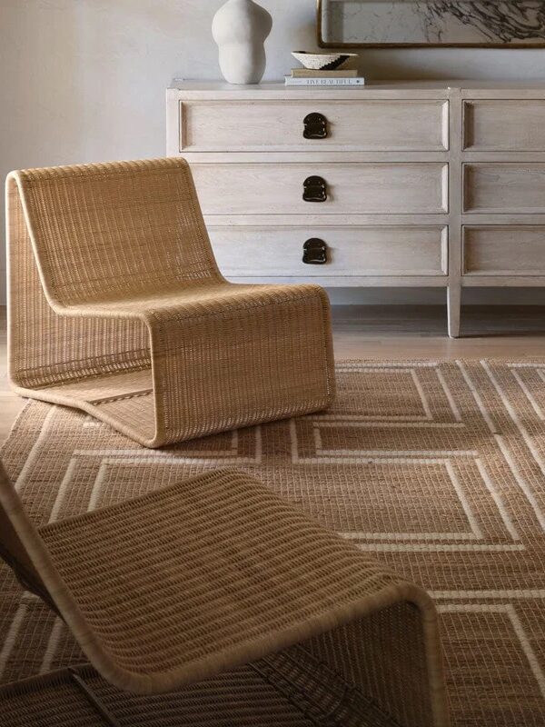 A minimalist room with woven chairs, a patterned rug, and a light wood dresser topped with a sculpture and mirror.
