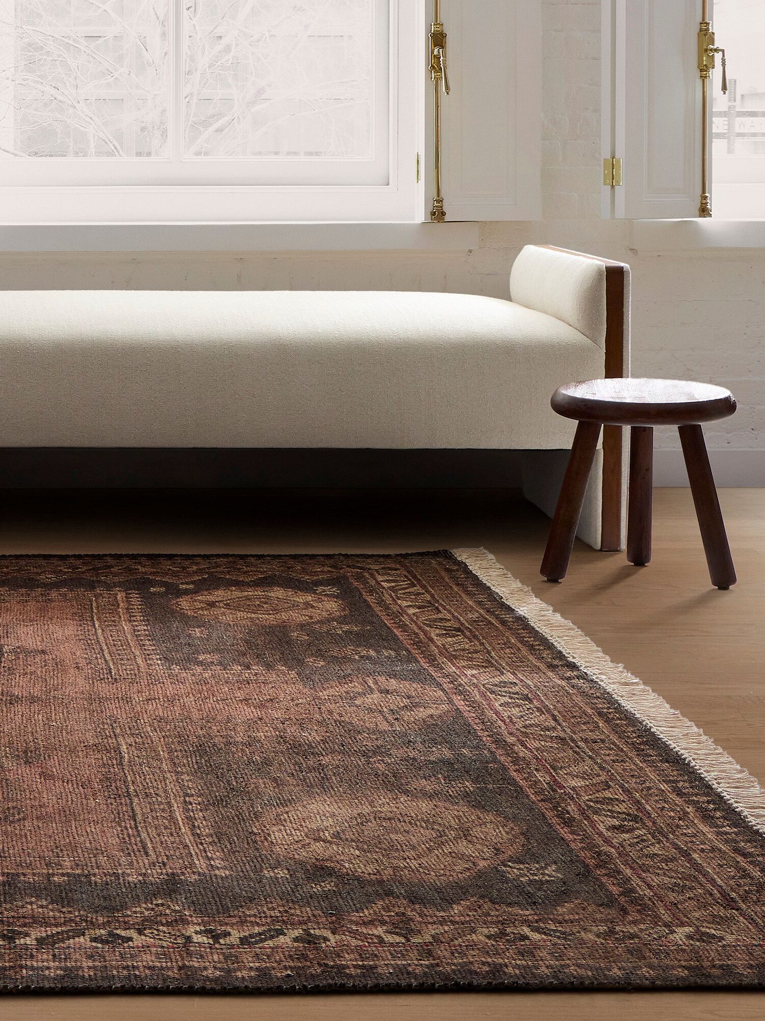 A minimalist room with a wooden bench, a round stool, and a patterned rug. A window with white frames and sheer curtains is in the background.