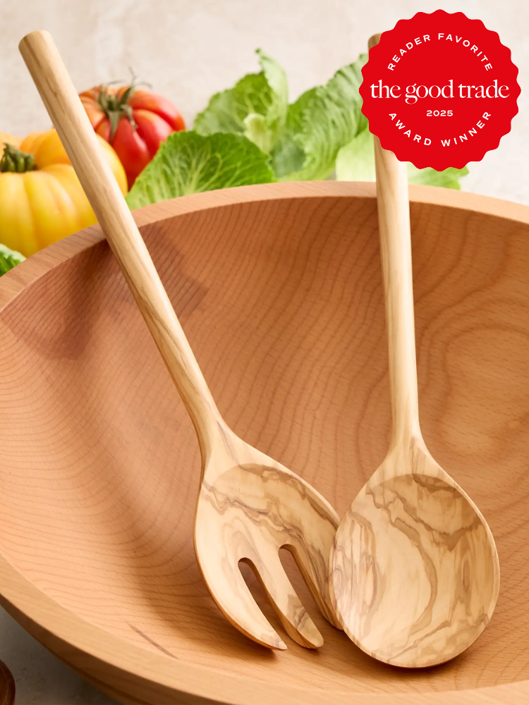 A wooden salad bowl with matching wooden serving fork and spoon, placed in front of fresh vegetables. Red award badge in upper right corner reads “The Good Trade Reader Favorite 2023 Award Winner.”.