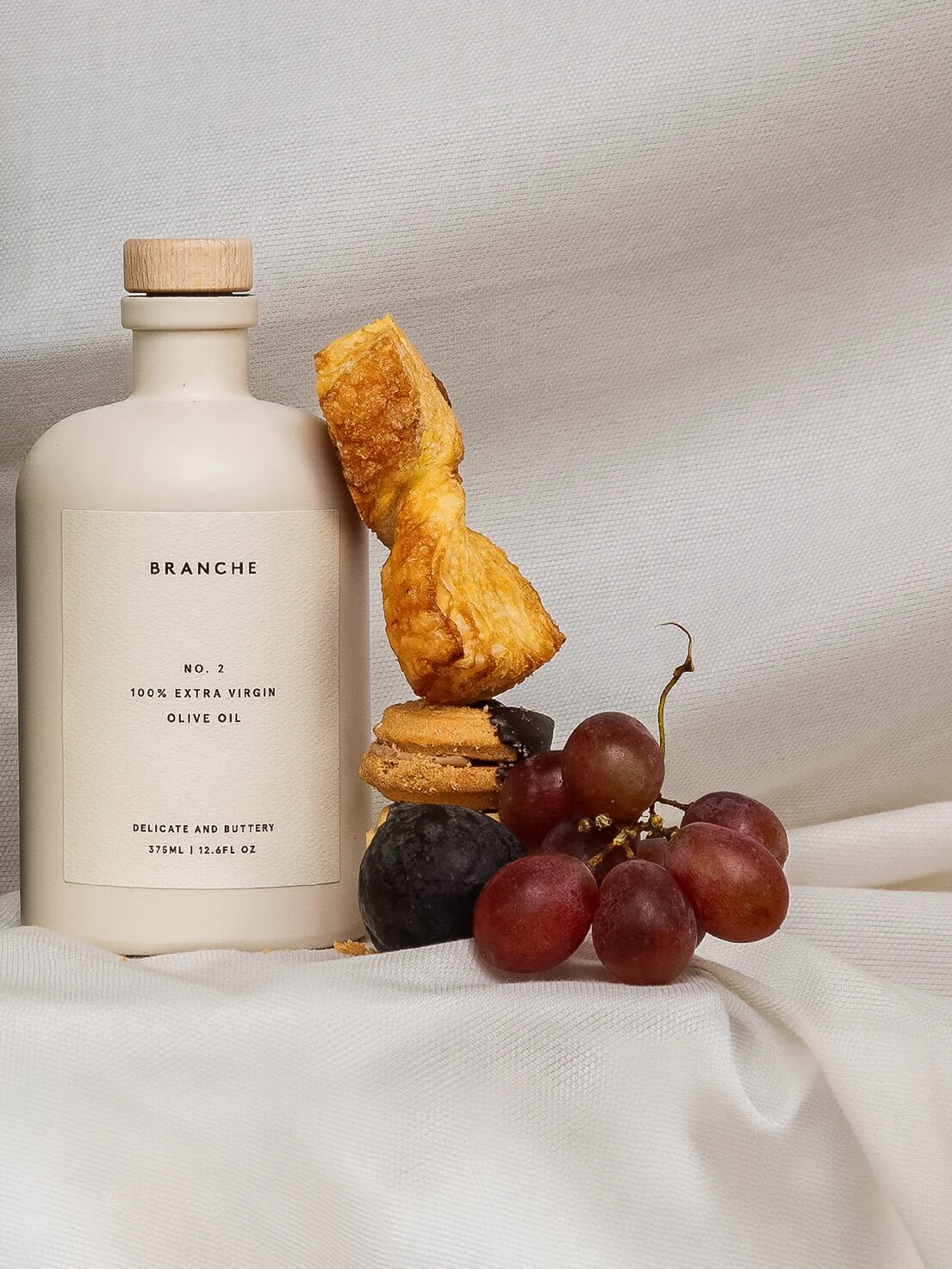 A bottle of olive oil is displayed beside stacked pastries, grapes, and a plum on a white cloth background.