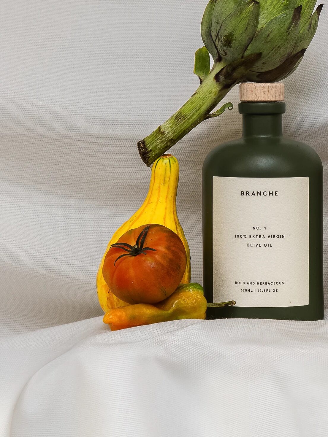 A bottle of olive oil labeled "Branche" sits beside an artichoke, a yellow squash, a tomato, and small peppers on a white fabric background.