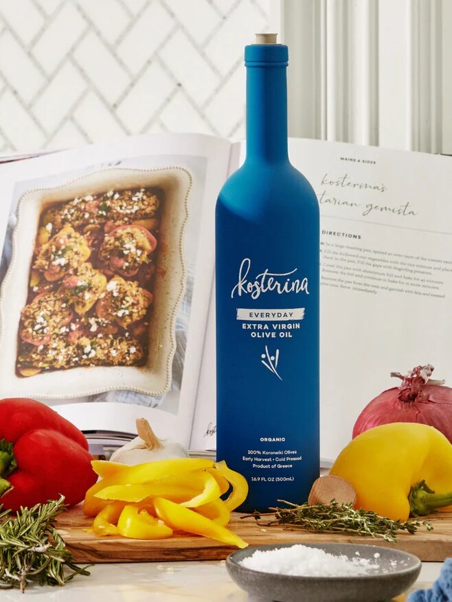 A bottle of Kosterina olive oil stands on a kitchen counter with bell peppers, herbs, and a cookbook.