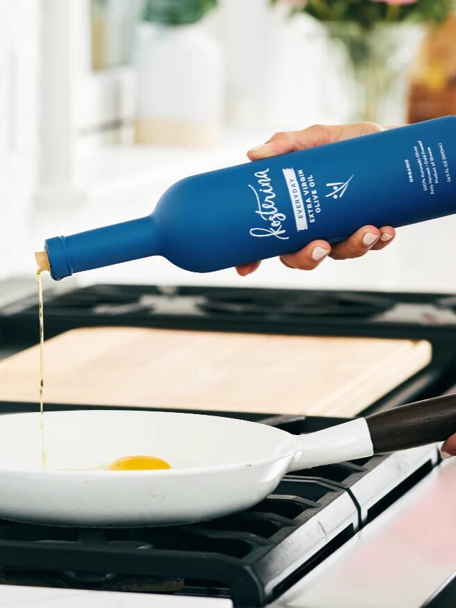 A hand pours olive oil from a blue bottle into a white frying pan with an egg on a stovetop.