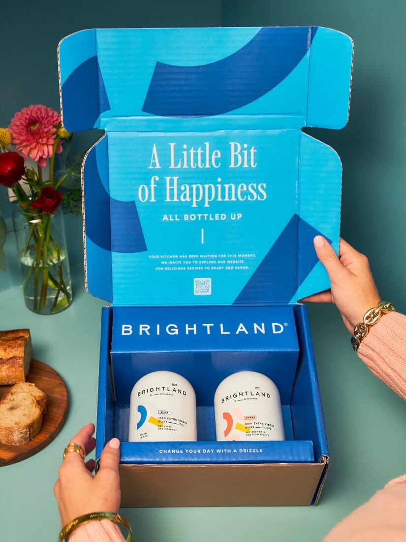 Person opening a blue Brightland box containing two bottles with text "A Little Bit of Happiness" on the lid. Bread, flowers, and a candle are in the background.