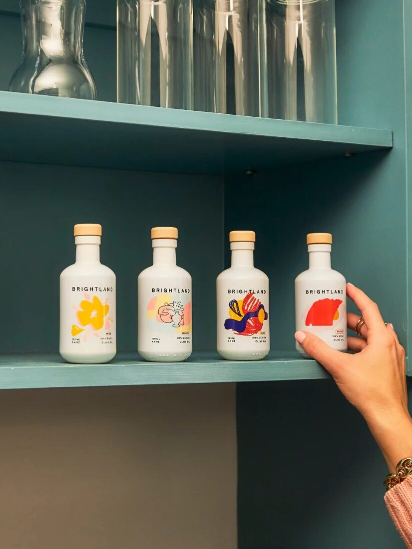Hand reaching for one of four brightly labeled bottles of olive oil on a blue kitchen shelf.