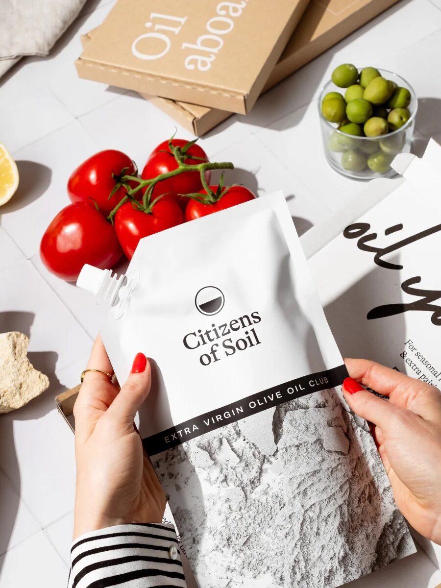 Person holding a pouch of "Citizens of Soil" extra virgin olive oil, surrounded by lemons, tomatoes, olives, baguette, and a bottle of wine on a white tiled surface.