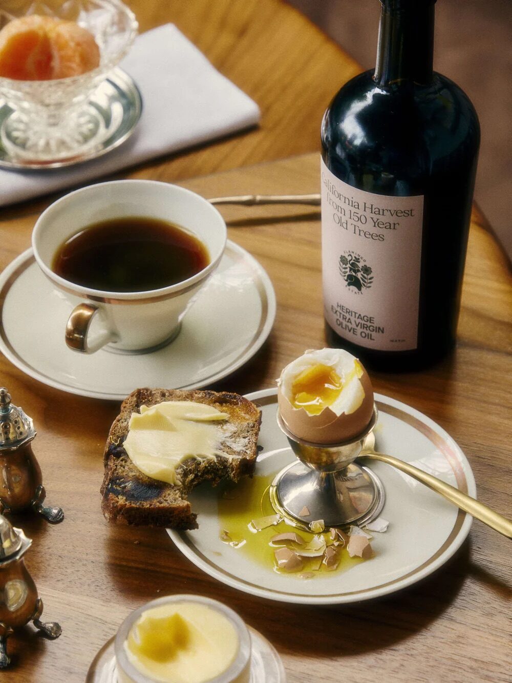 A breakfast table with coffee, a bottle of olive oil, buttered toast, a soft-boiled egg in an egg cup, and a small dish of butter on a wooden surface.