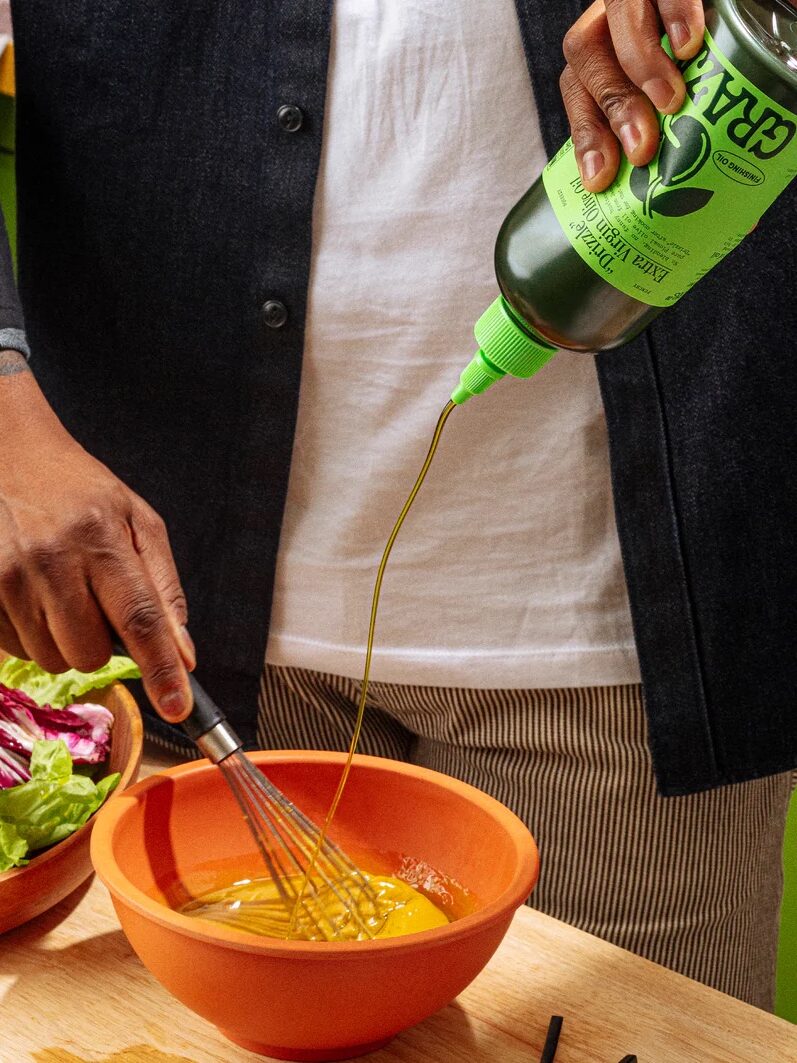 Person whisking in a bowl while pouring olive oil, with a salad bowl nearby on a wooden surface.