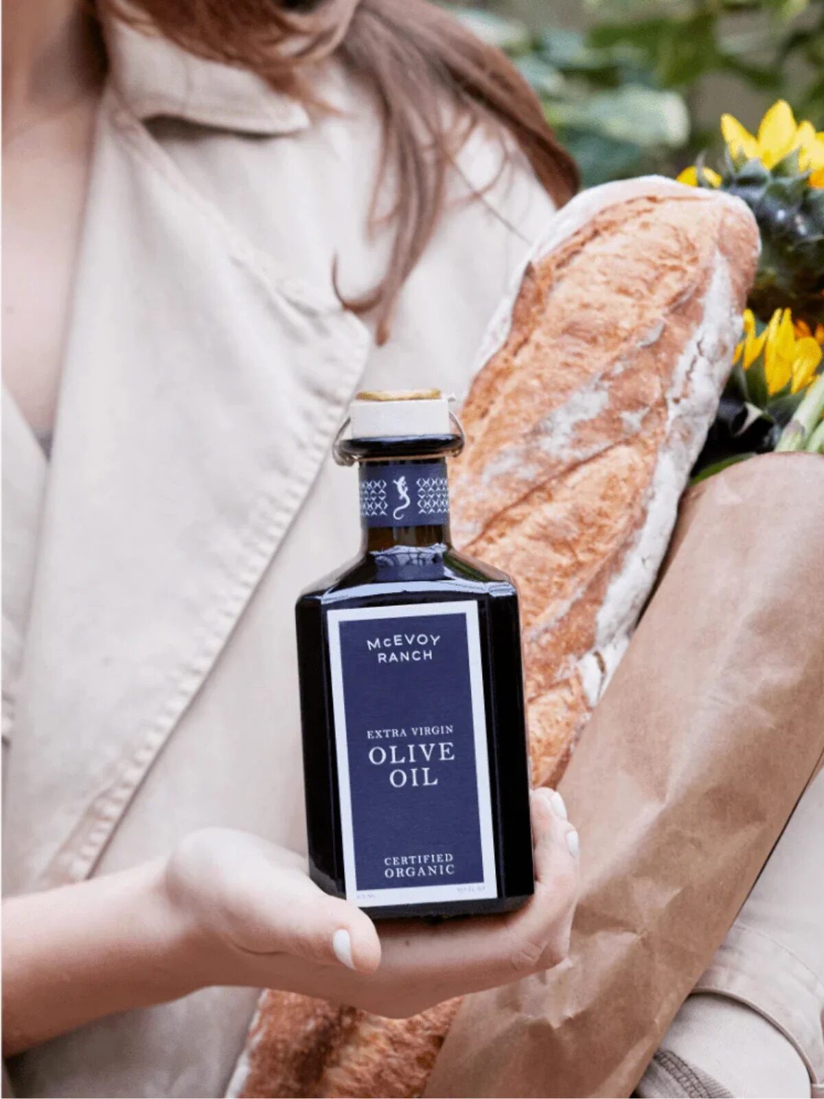 Person holding a bottle of olive oil, a baguette, and a bouquet of sunflowers wrapped in brown paper.