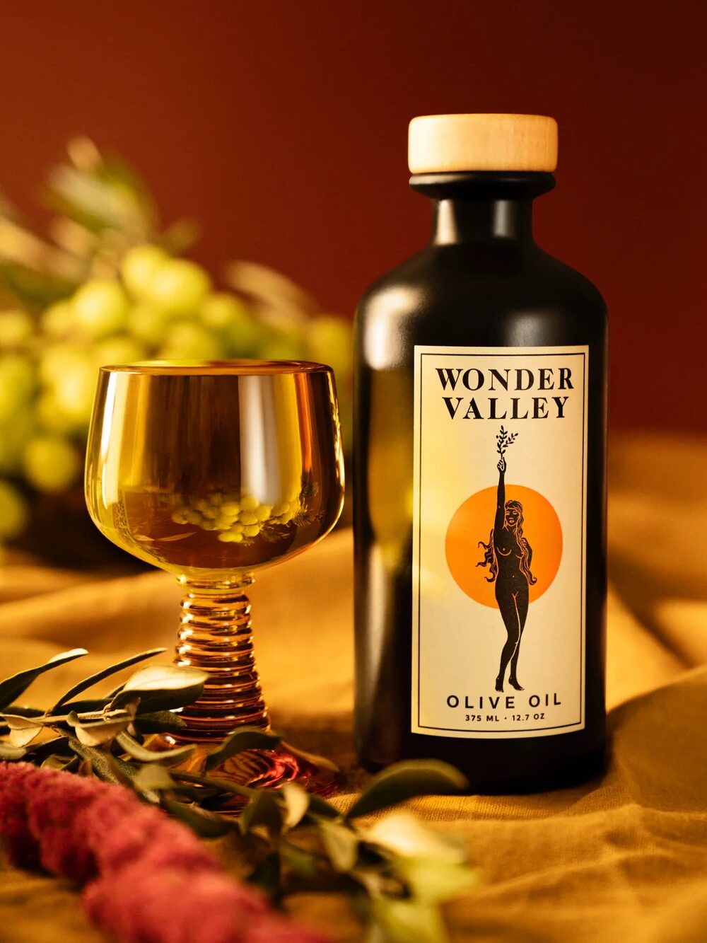 A bottle of Wonder Valley olive oil next to a gold-rimmed glass, with green olives in the background on a yellow surface.
