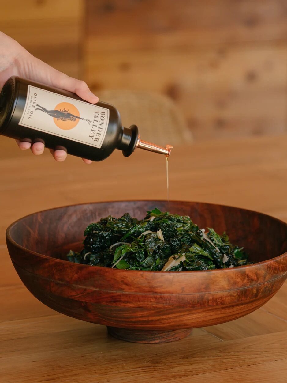 Hand pouring olive oil from a bottle onto a bowl of kale salad on a wooden table.