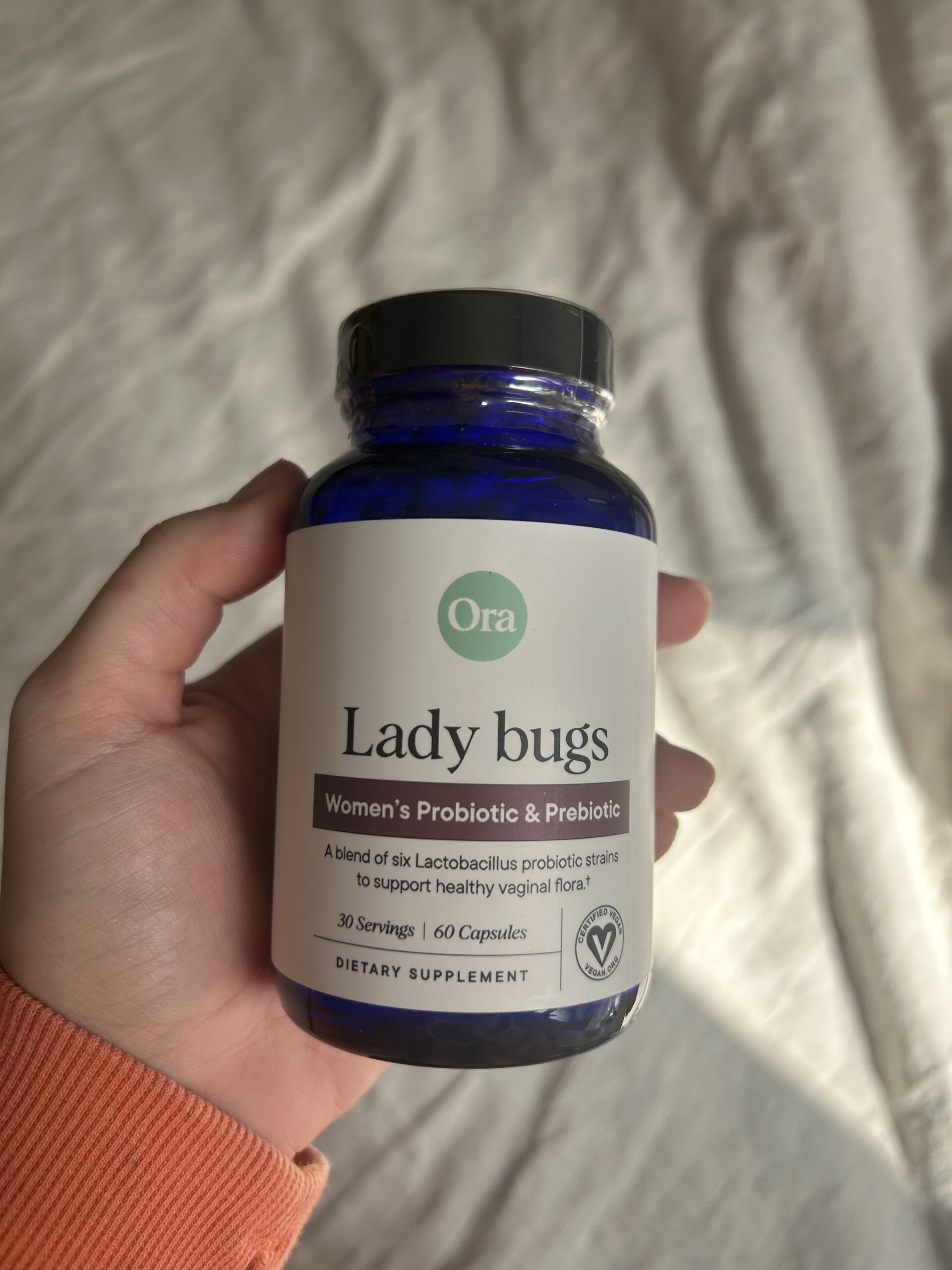 A hand holds a blue bottle labeled "Lady bugs Women's Probiotic & Prebiotic" by Ora, containing 60 capsules for vaginal health support.