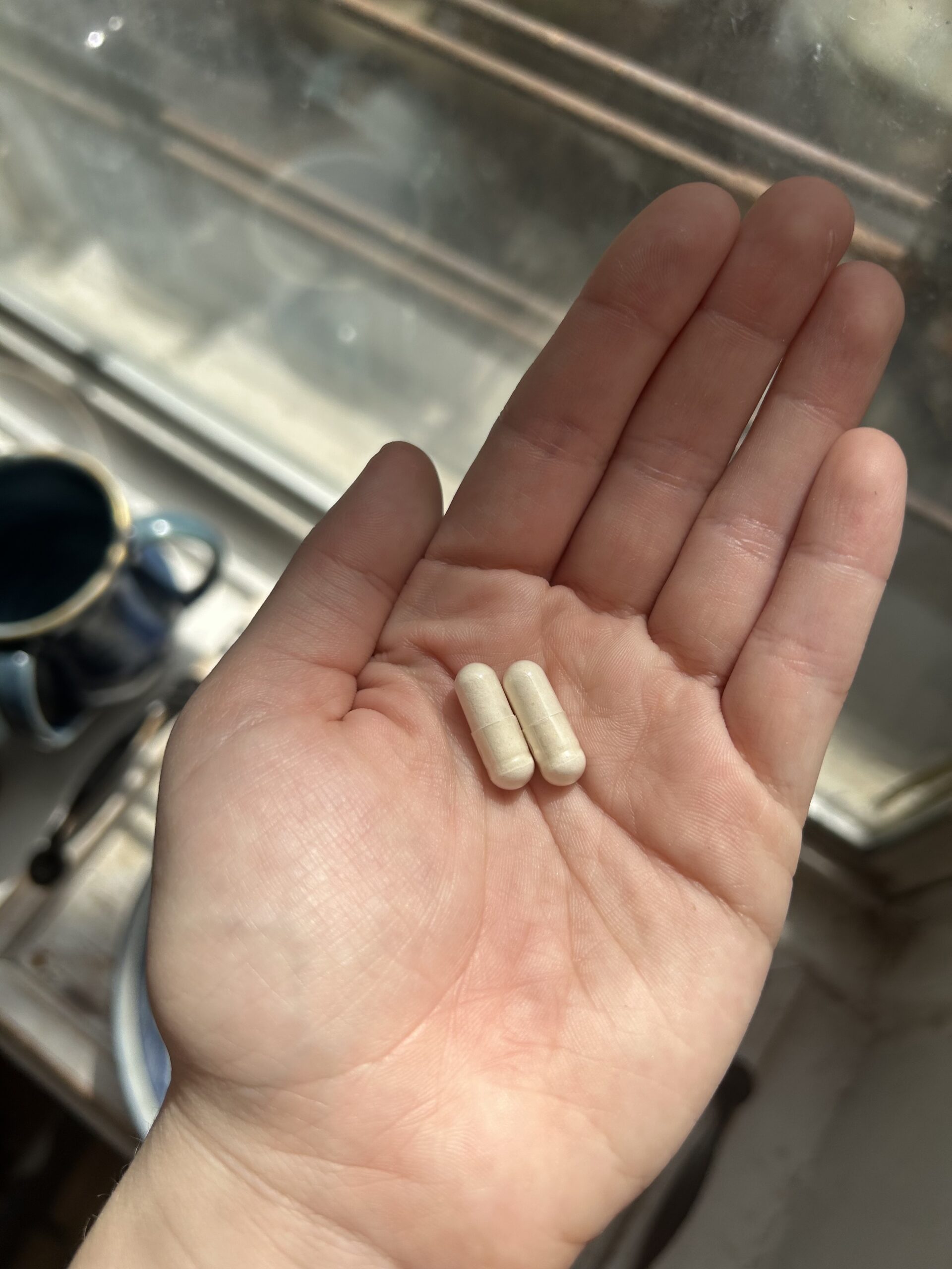 A hand holding two white capsules with a window and cup visible in the background.