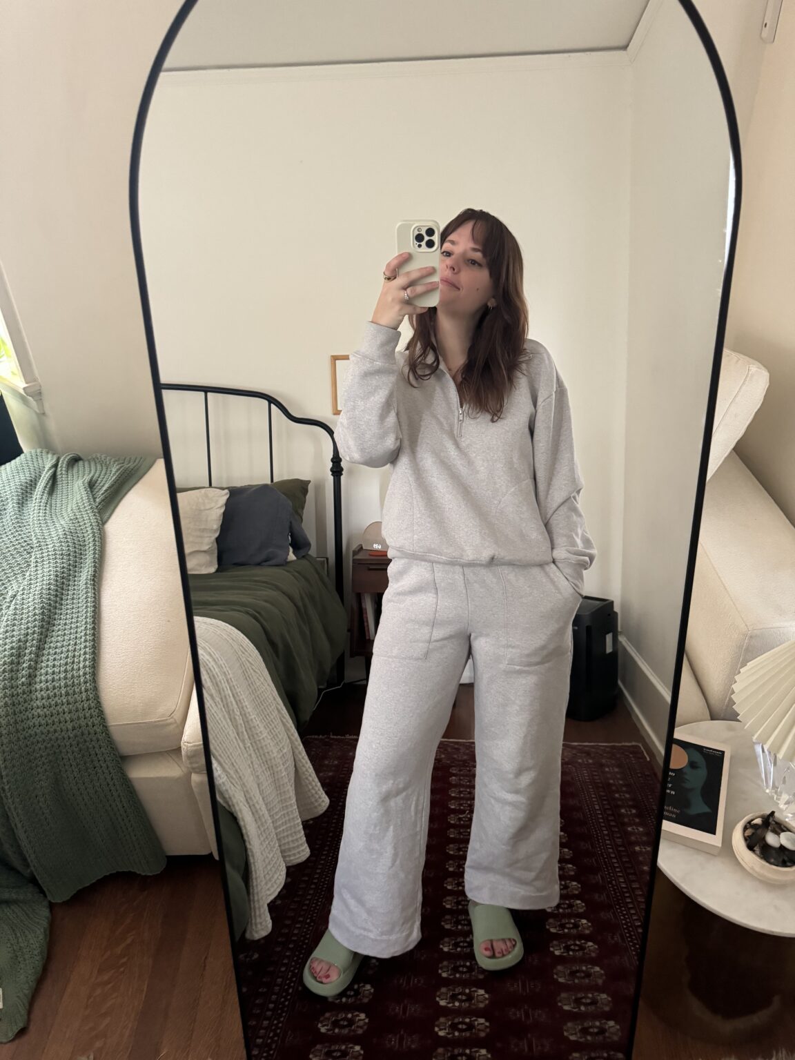 Person in a gray tracksuit taking a mirror selfie in a room with a bed, green blanket, and a small side table.