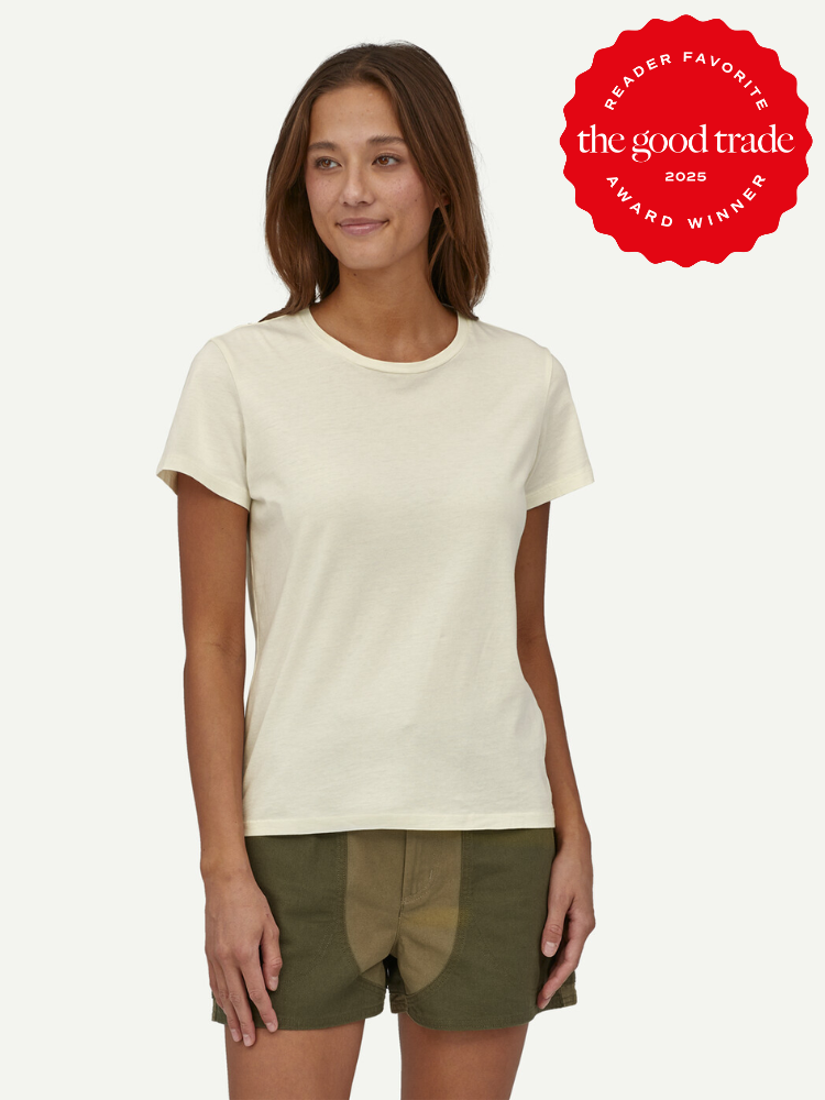 Woman wearing a cream-colored short-sleeve t-shirt and green shorts stands against a plain background; a red "The Good Trade 2025 Award Winner" badge appears in the corner.