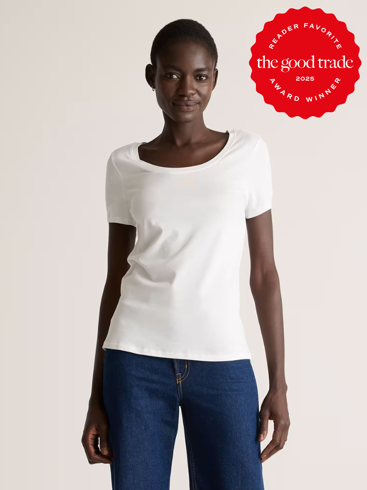 A woman wearing a plain white short-sleeve t-shirt and blue jeans stands against a neutral background. A red award badge in the corner reads "The Good Trade Reader Favorite 2025 Award Winner.