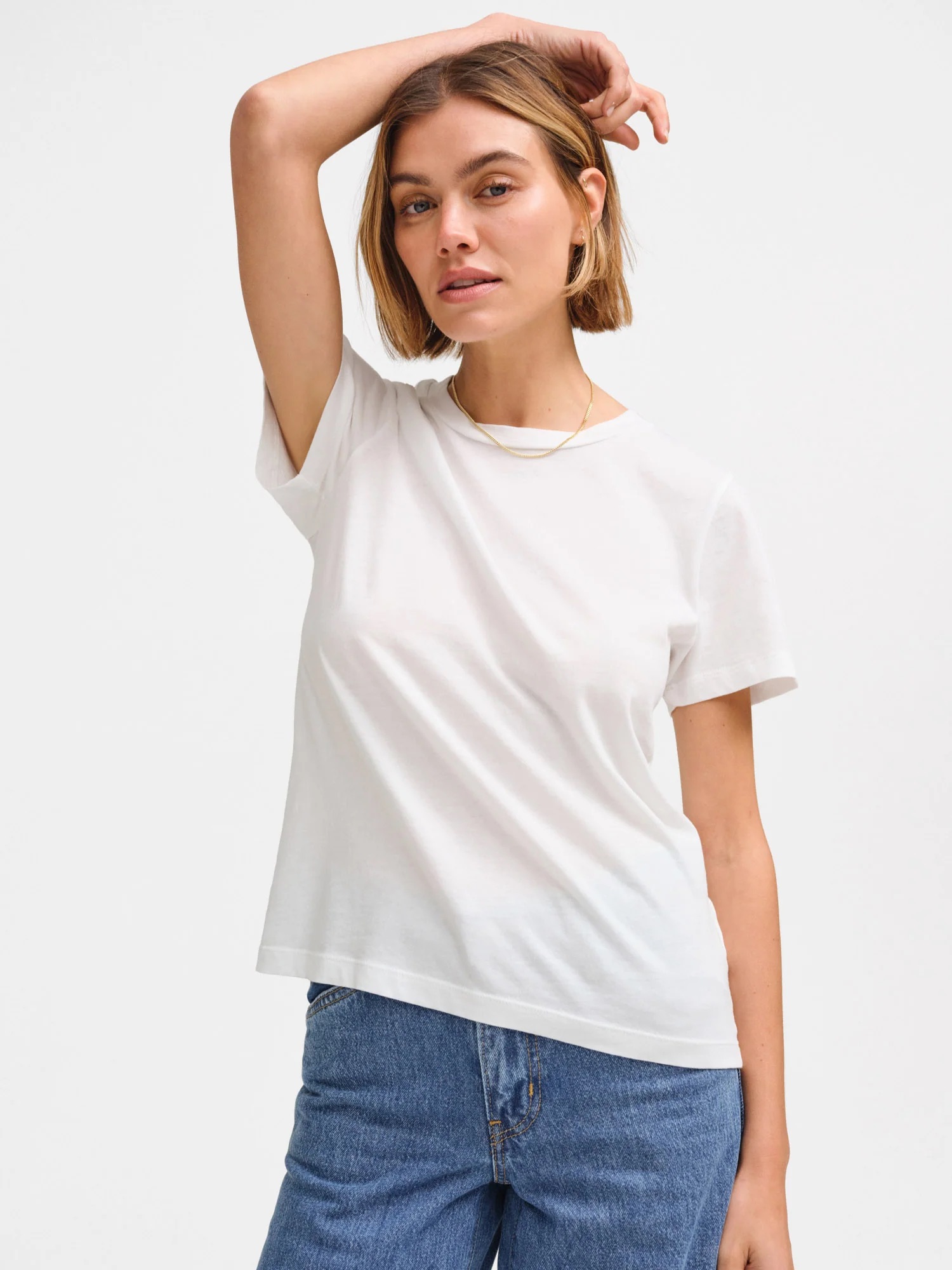 A woman with short hair wears a plain white t-shirt and blue jeans, standing with one arm raised and touching her head against a plain background.