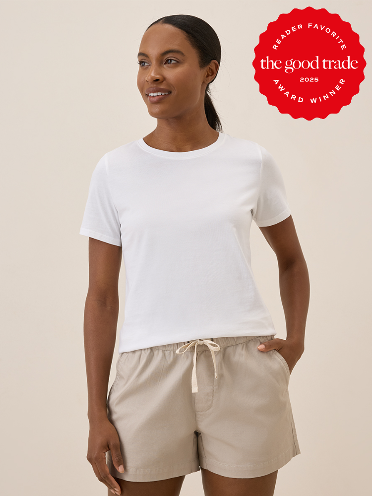 A woman wears a plain white t-shirt and beige drawstring shorts, standing against a neutral background. A red "The Good Trade Award Winner 2025" badge appears in the upper right corner.